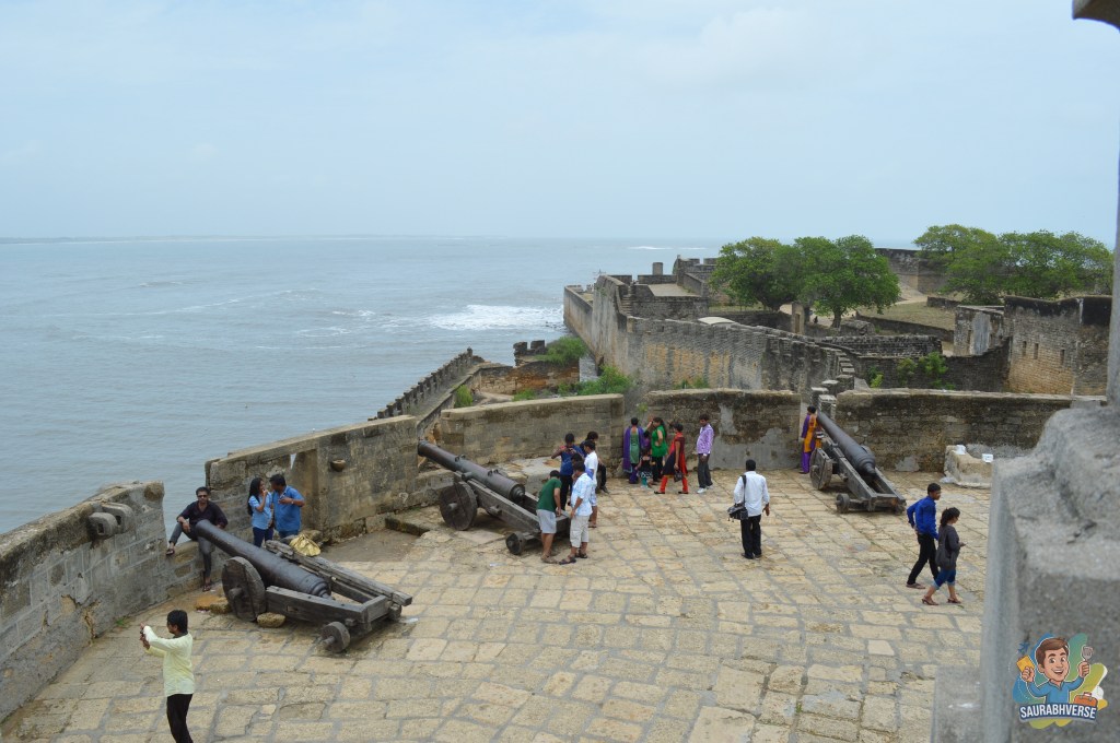 A panoramic view of the Diu Fort featuring historical cannons, visitors exploring the area, and the Arabian Sea in the background under a cloudy sky.